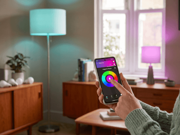 Somebody holding a smart phone and pressing the screen to change the colour of LED smart bulbs in lamps in a living room.