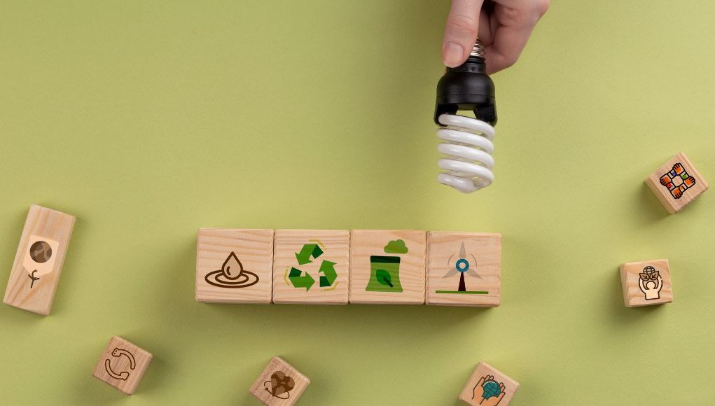 Hand holding a CFL lamp over some blocks with different symbols related to recycling.
