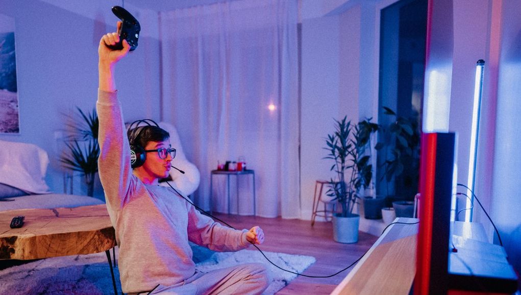 A man celebrating while holding a game console controller in a room lit with a blue and red light.
