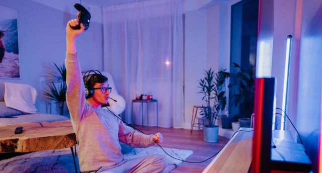 A man celebrating while holding a game console controller in a room lit with a blue and red light.
