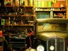 A garage interior basked in warm light and filled with a lot of trinkets and an old car on one side.