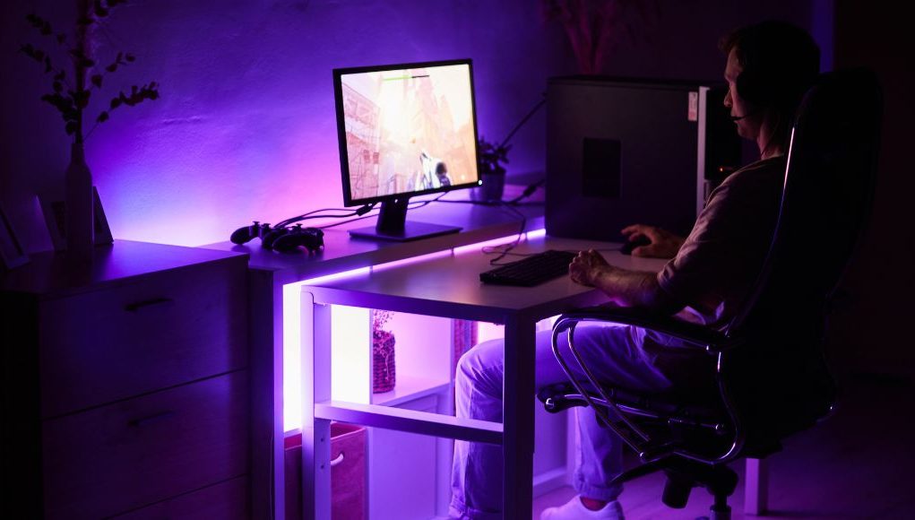 A man sitting in front of a PC in a dark room with an under desk purple light.