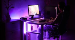 A man sitting in front of a PC in a dark room with an under desk purple light.