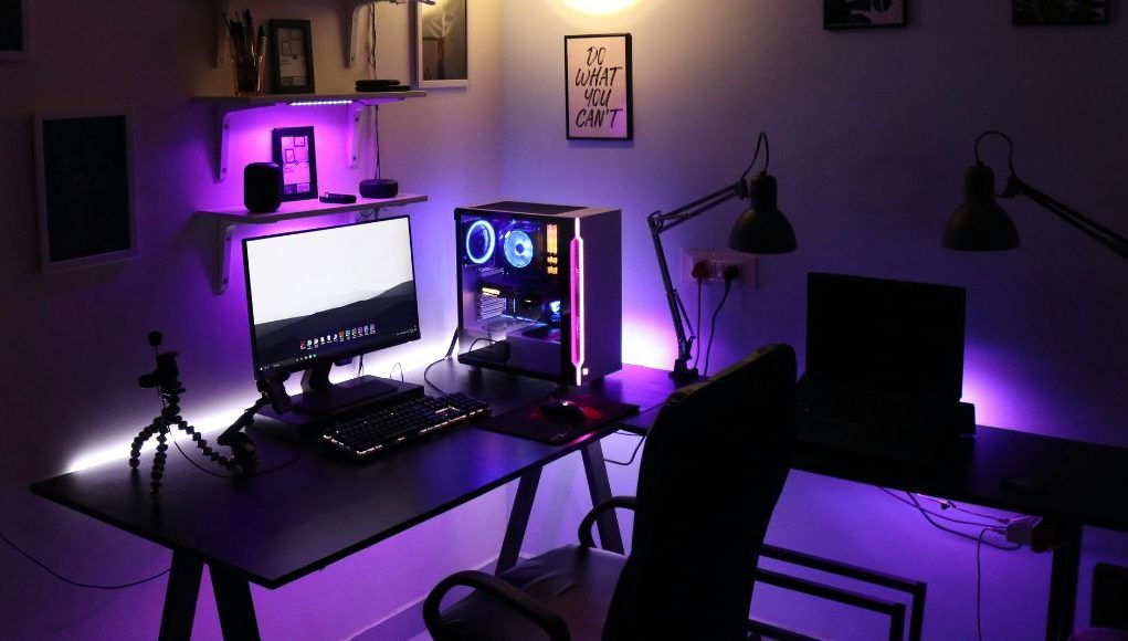 A PC setup in a dim room with a warm overhead light and a purple under desk lighting.