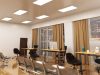An open office space with three desks near two big glass windows and multiple LED panel lights on the ceiling.