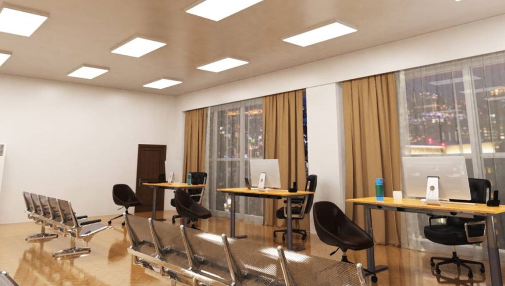 An open office space with three desks near two big glass windows and multiple LED panel lights on the ceiling.
