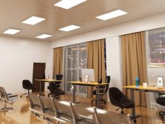 An open office space with three desks near two big glass windows and multiple LED panel lights on the ceiling.