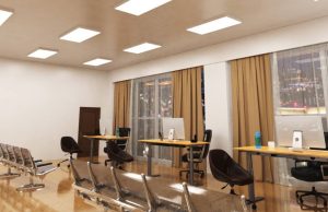 An open office space with three desks near two big glass windows and multiple LED panel lights on the ceiling.