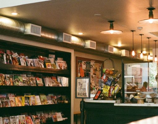 An view of a commercial space from the outside with shelves, the counter, and warm lights visible.