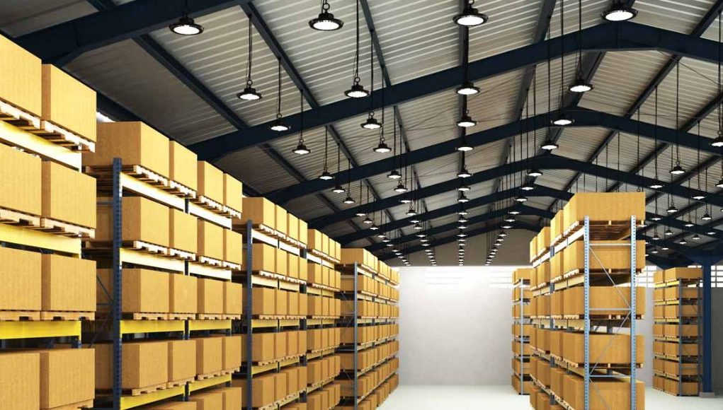 An image of a warehouse with shelves filled with boxes and the ceiling filled with high bay lights.