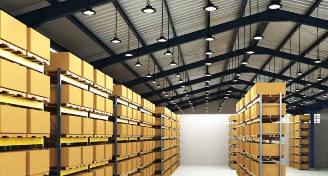 Industrial & Warehouse Lighting Strategies for High Ceilings An image of a warehouse with shelves filled with boxes and the ceiling filled with high bay lights.