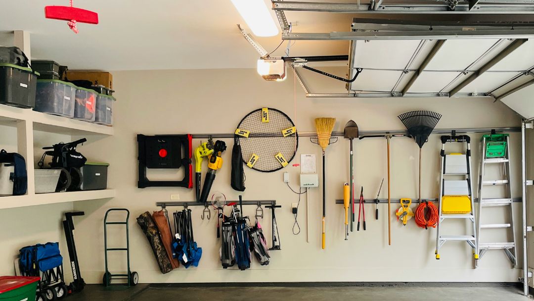 A garage wall with multiple tools hanging on it, a single light shining on the ceiling.