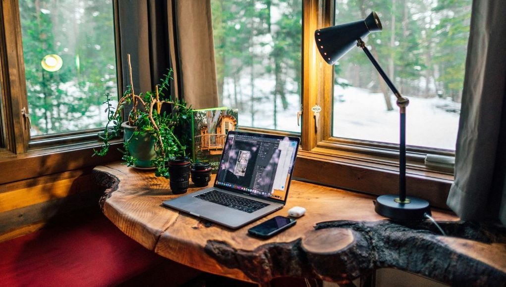 A wooden desk stationed by the window with a desk lamp glowing with warm light beside it.
