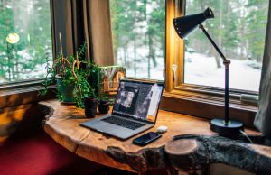 A wooden desk stationed by the window with a desk lamp glowing with warm light beside it.