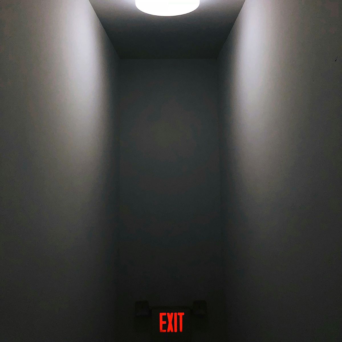 An exit hallways with grey walls, a bright white light above, and a red exit sign at the bottom of the image.
