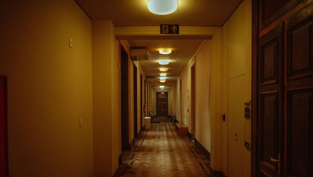 Commercial vs. Residential Emergency Lighting hero image A residential building hallway with warm lighting and a visible emergency exit sign on one of the beams.