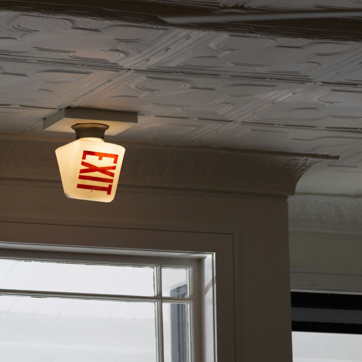 A white exit sign with red text and a warm glow installed on a patterned ceiling near a window that lets in natural light during the day.