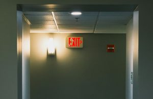 Emergency Lighting for Small Businesses A corridor wall with a red EXIT sign beside a wall light with a warm glow.