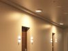 Self-Test Emergency Lights Explained A hallway with multiple doors illuminated by round bulkhead lights on the ceiling and wall lights on both sides of each door.