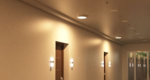 A hallway with multiple doors illuminated by round bulkhead lights on the ceiling and wall lights on both sides of each door.