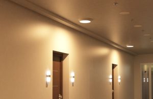 Self-Test Emergency Lights Explained A hallway with multiple doors illuminated by round bulkhead lights on the ceiling and wall lights on both sides of each door.