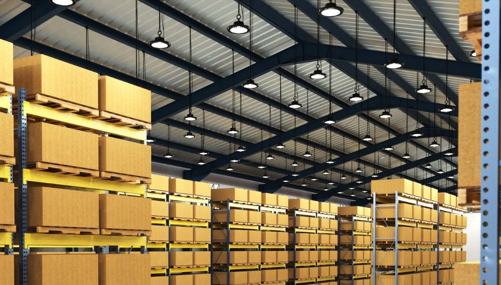 A warehouse with multiple boxes on racks and multiple high bay lights hanging from the ceiling.