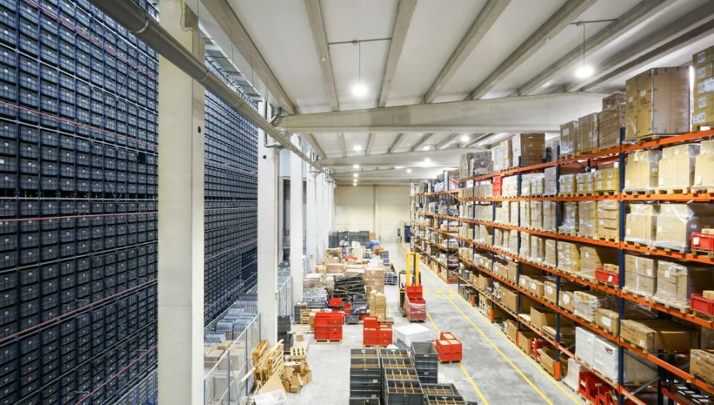 A large warehouse with multiple rack filled with boxes, the ground is also filled with large items and forklifts.