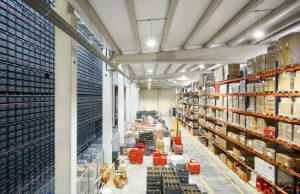 A large warehouse with multiple rack filled with boxes, the ground is also filled with large items and forklifts.