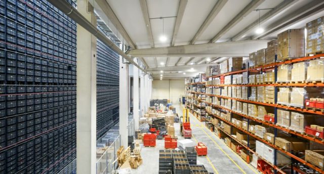A large warehouse with multiple rack filled with boxes, the ground is also filled with large items and forklifts.