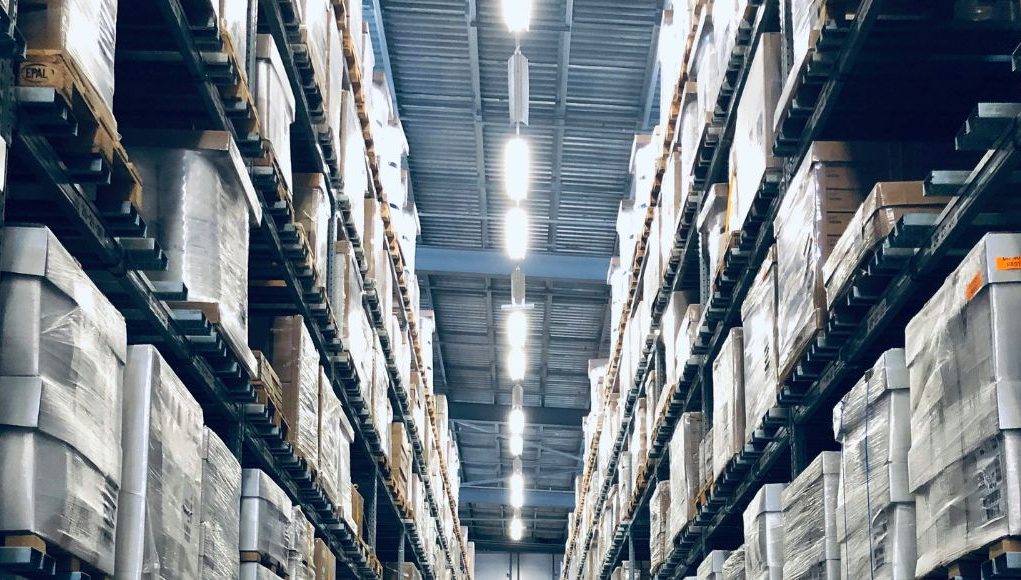 A warehouse aisle with racks filled from top to bottom with boxes, at the middle on the roof are multiple bright white light sources illuminating the space below.