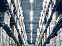 A warehouse aisle with racks filled from top to bottom with boxes, at the middle on the roof are multiple bright white light sources illuminating the space below.