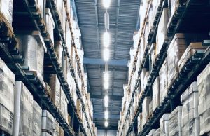 Retrofit vs. New Installation: Upgrading Warehouse Lighting Systems A warehouse aisle with racks filled from top to bottom with boxes, at the middle on the roof are multiple bright white light sources illuminating the space below.