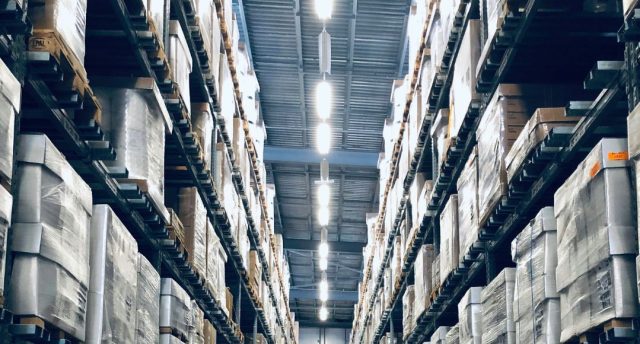 A warehouse aisle with racks filled from top to bottom with boxes, at the middle on the roof are multiple bright white light sources illuminating the space below.