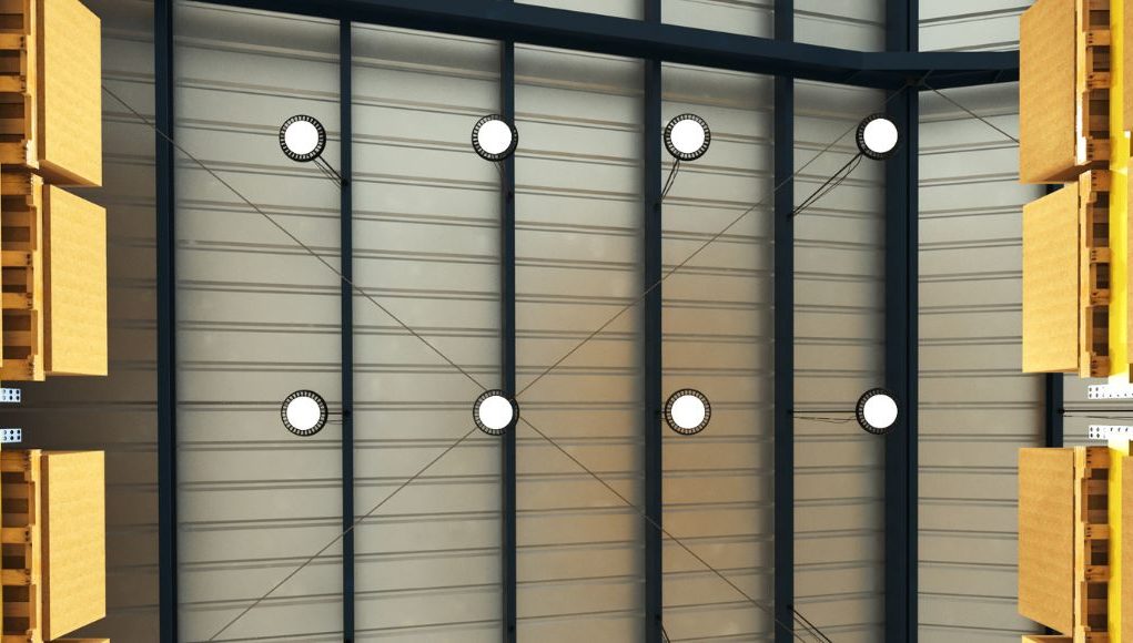A ware house ceiling lined with multiple high bay lights with white glow, boxes on racks to the side.