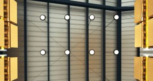 A ware house ceiling lined with multiple high bay lights with white glow, boxes on racks to the side.