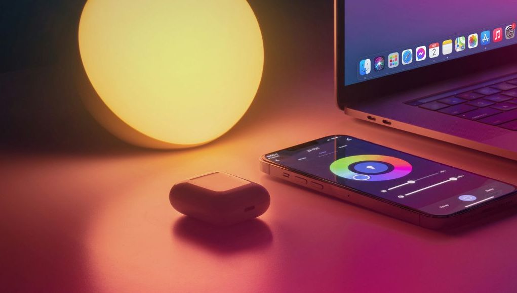 A warm light, airpods, laptop, and phone with colour controls for the light on a desk.