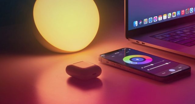 A warm light, airpods, laptop, and phone with colour controls for the light on a desk.