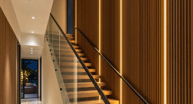 A modern home staircase with slat wall panel on one side with multiple grooves illuminated by warm white LED neon slat lighting.